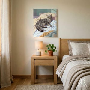 May include: A square wall art print featuring a digital illustration of a grey tabby cat resting on rumpled white and yellow bedding. The cat is positioned in the center of the composition, looking forward with alert eyes against a soft teal background.