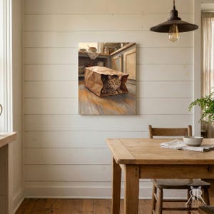 May include: A rectangular wall art print depicting a brown tabby cat sitting inside a tan paper grocery bag. The scene is set on a wooden floor within a kitchen environment featuring light-colored cabinets and shelves in the background.