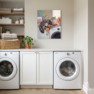 May include: A wall art print depicting a fluffy grey and black tabby cat resting on a pile of colorful laundry atop a white washing machine. The scene includes stacked towels on shelves, a woven basket, and a small potted plant in a laundry room.