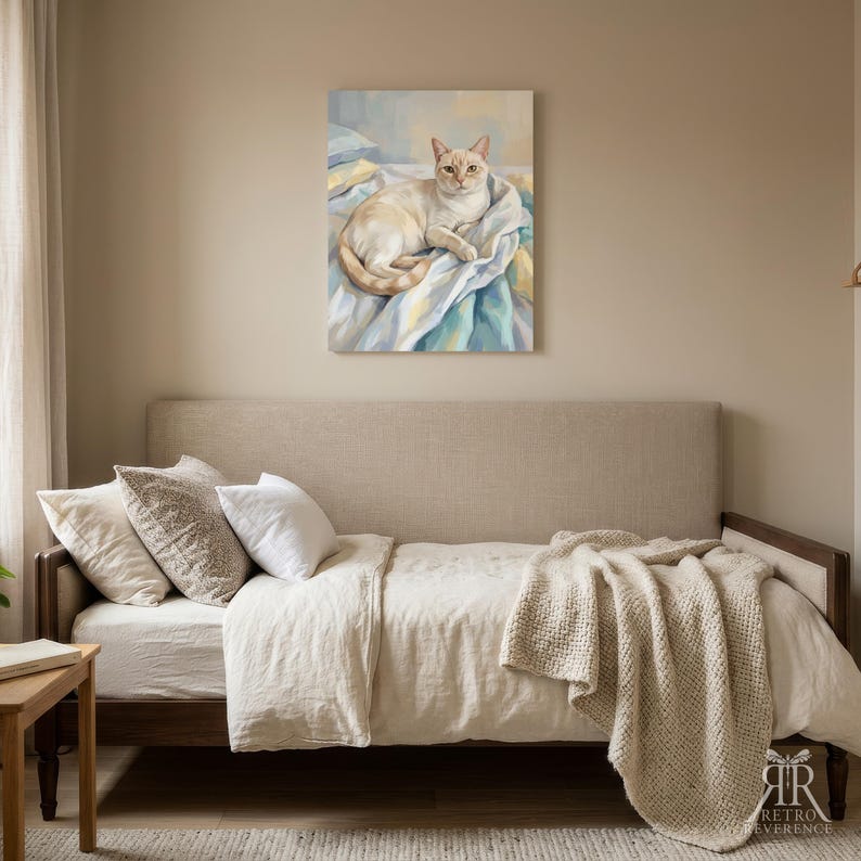May include: A rectangular wall art print featuring a painting of a cream-colored cat with pointed ears and blue eyes, resting on a rumpled bed with blue, yellow, and white linens. The artwork is displayed on a plain beige wall above a daybed.