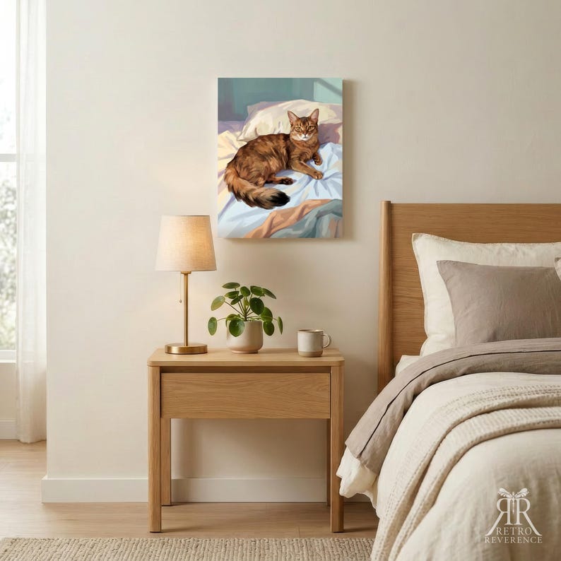 May include: A rectangular wall art print featuring a brown tabby cat resting on white and blue bedding. The artwork is displayed above a light wood nightstand that holds a lamp with a beige shade, a small green potted plant, and a ceramic mug.