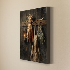 May include: A square canvas wall art print featuring a dark, weathered wooden wall with a horizontal plank holding a metal lantern, a bundle of dried orange corn cobs, a beige fabric apron, and a bunch of dried green herbs hanging from hooks.