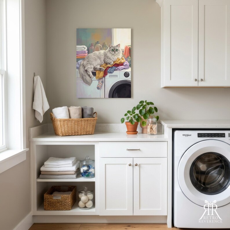 May include: A wall art print depicting a fluffy white cat resting on top of a white washing machine amidst a pile of colorful laundry. The cat has long fur and green eyes. The artwork is displayed in a laundry room with white cabinets and baskets.
