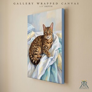 May include: A gallery wrapped canvas with a 1.5 inch depth featuring a painting of a brown spotted Bengal cat resting on white and light blue rumpled bedding. The artwork displays the cat in a seated position with its tail curled.