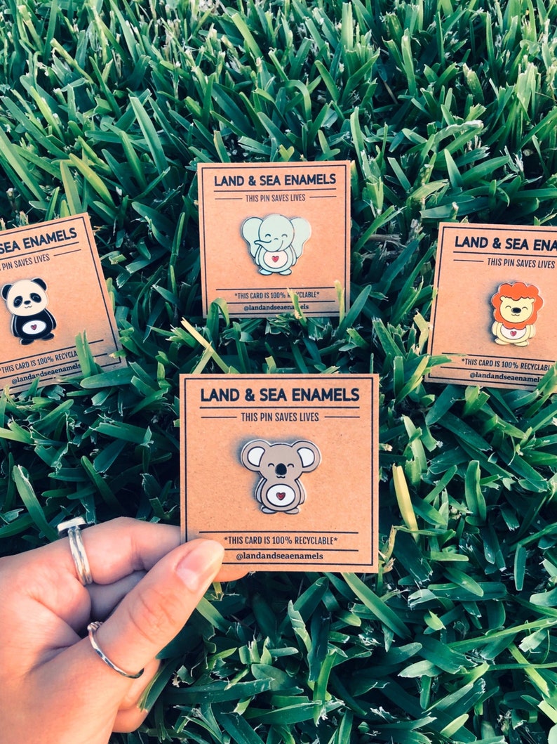 May include: Three brown cardstock cards with white text and colorful enamel pins. Each card features a different animal: an elephant, a koala, and a lion. The text on the cards reads "Land & Sea Enamels" and "This pin saves lives". The cards also include the text "This card is 100% recyclable" and "@landandseaenamels".