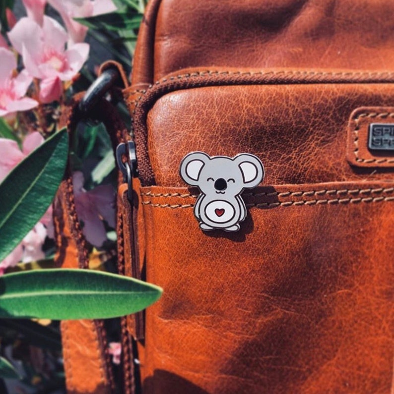May include: A gray koala enamel pin with a red heart on a brown leather bag.