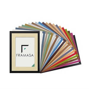 May include: A black rectangular picture frame with a cream-coloured mount containing the text FRAMASA in black. Behind the frame, a fan-shaped display of numerous picture frame samples in various colours, textures, and wood grain finishes.