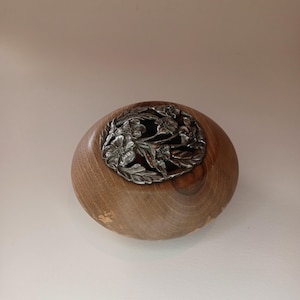May include: A wooden bowl with a silver floral design on top. The bowl is a light brown color and has a smooth, rounded shape. The silver design is intricate and detailed, featuring flowers and leaves.