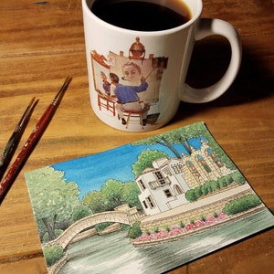May include: A white ceramic mug filled with dark coffee, featuring a painting of an artist at work. Beside the mug are two paintbrushes and a watercolor painting of a building with a bridge over water. The scene is set on a wooden surface.