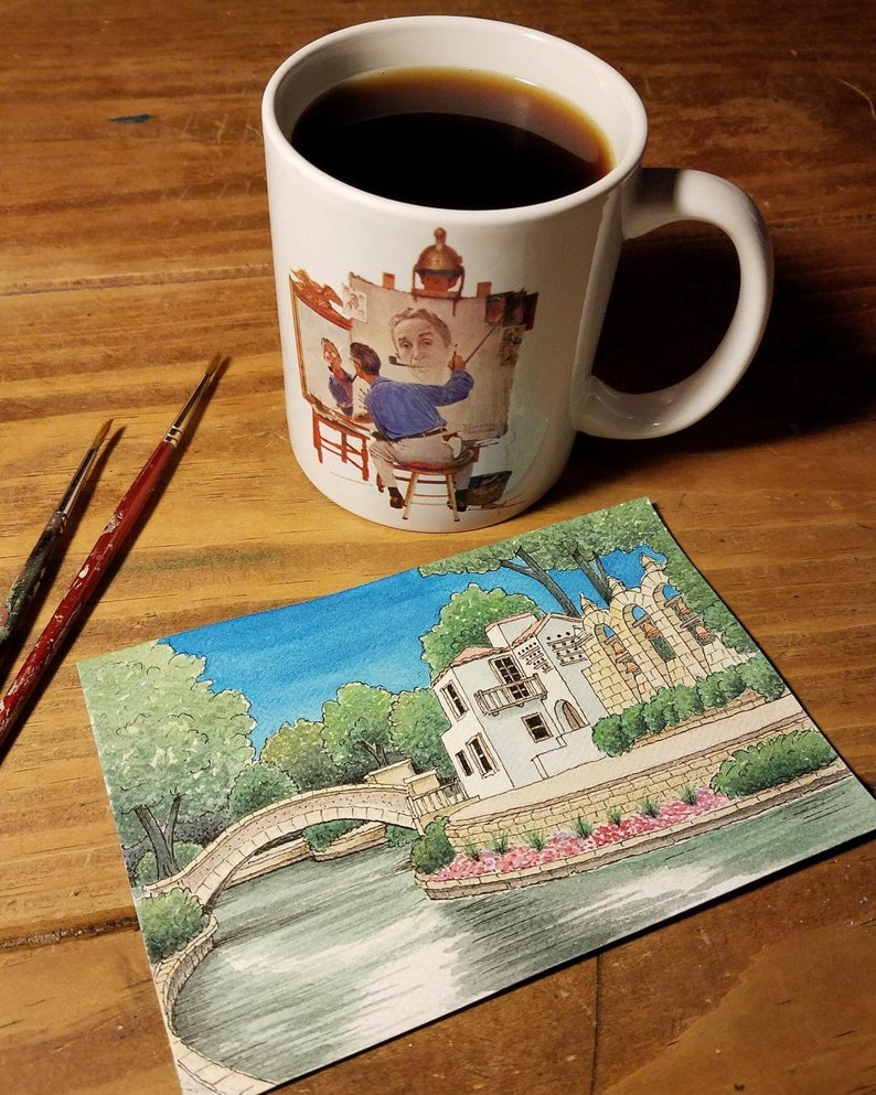 May include: A white ceramic mug filled with dark coffee, featuring a painting of an artist at work. Beside the mug are two paintbrushes and a watercolor painting of a building with a bridge over water. The scene is set on a wooden surface.