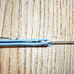 May include: A light blue folding pocket knife with a silver blade. The knife is open, revealing the blade. The handle has a pocket clip. The knife is on a light brown wooden surface.