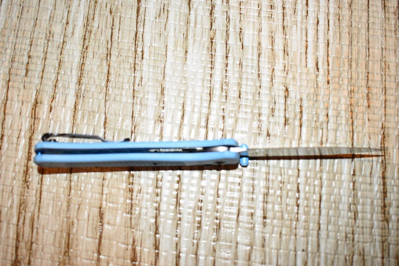May include: A light blue folding pocket knife with a silver blade. The knife is open, revealing the blade. The handle has a pocket clip. The knife is on a light brown wooden surface.