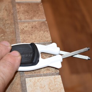 May include: A white and black handle tool with two silver metal rods extending from it. The tool is held over a brown and tan tiled surface. The handle has a black, textured grip.
