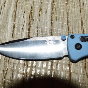 May include: A close-up of a folding knife with a silver blade and a light blue handle. The blade has a black edge and a butterfly logo. The handle has a textured surface and black hardware.