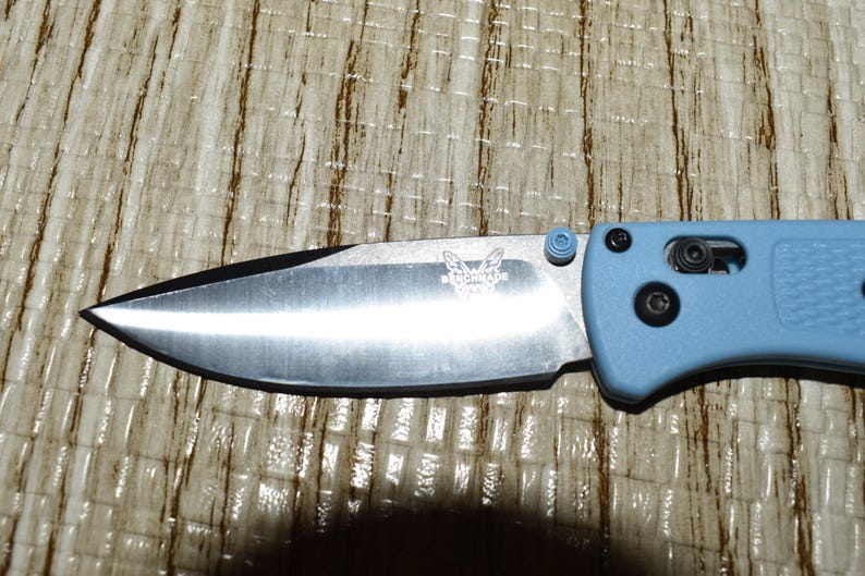 May include: A close-up of a folding knife with a silver blade and a light blue handle. The blade has a black edge and a butterfly logo. The handle has a textured surface and black hardware.