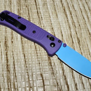 May include: A purple folding knife with a light blue blade. The handle has a textured surface and a black pocket clip. The knife is closed and resting on a textured surface.
