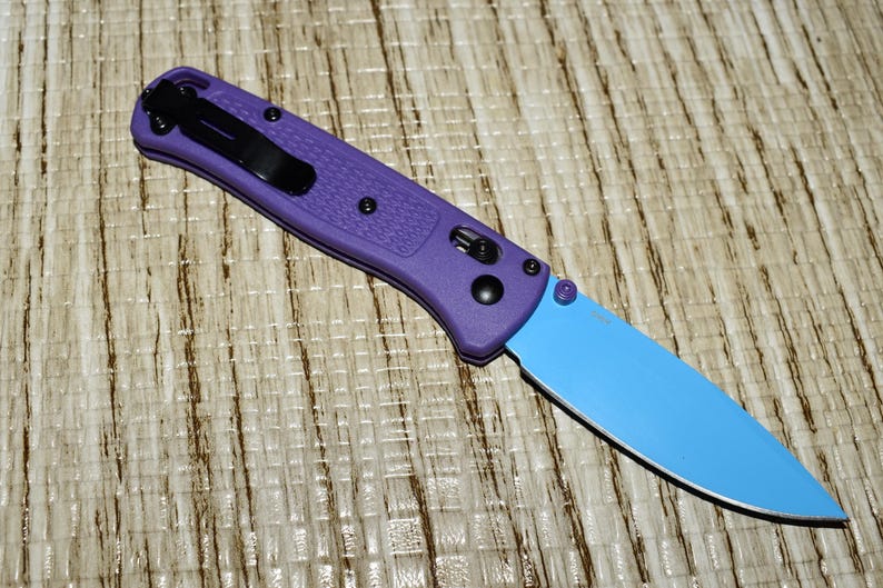 May include: A purple folding knife with a light blue blade. The handle has a textured surface and a black pocket clip. The knife is closed and resting on a textured surface.
