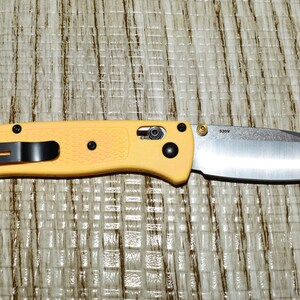 May include: A yellow folding knife with a silver blade. The knife has a black pocket clip and black hardware. The blade is partially open, and the handle is textured. The knife is resting on a textured surface.