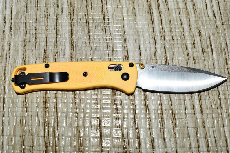 May include: A yellow folding knife with a silver blade. The knife has a black pocket clip and black hardware. The blade is partially open, and the handle is textured. The knife is resting on a textured surface.