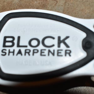 May include: Close-up of a white and black knife sharpener. The text "BLOCK SHARPENER" is printed in bold black letters on the white surface. The sharpener has a black rubber ring around the center.