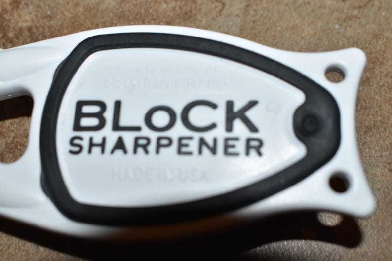May include: Close-up of a white and black knife sharpener. The text "BLOCK SHARPENER" is printed in bold black letters on the white surface. The sharpener has a black rubber ring around the center.