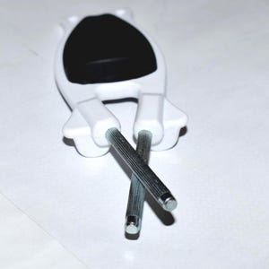 May include: A white plastic cabinet door handle with a black oval center and two silver metal rods extending downwards. The rods are crossed. The handle is designed to be mounted on a cabinet door.