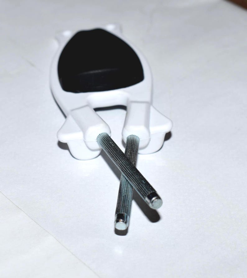 May include: A white plastic cabinet door handle with a black oval center and two silver metal rods extending downwards. The rods are crossed. The handle is designed to be mounted on a cabinet door.