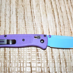 May include: A purple folding knife with a blue blade. The knife has a black pocket clip and black hardware. The handle is textured, and the blade is a solid blue color.