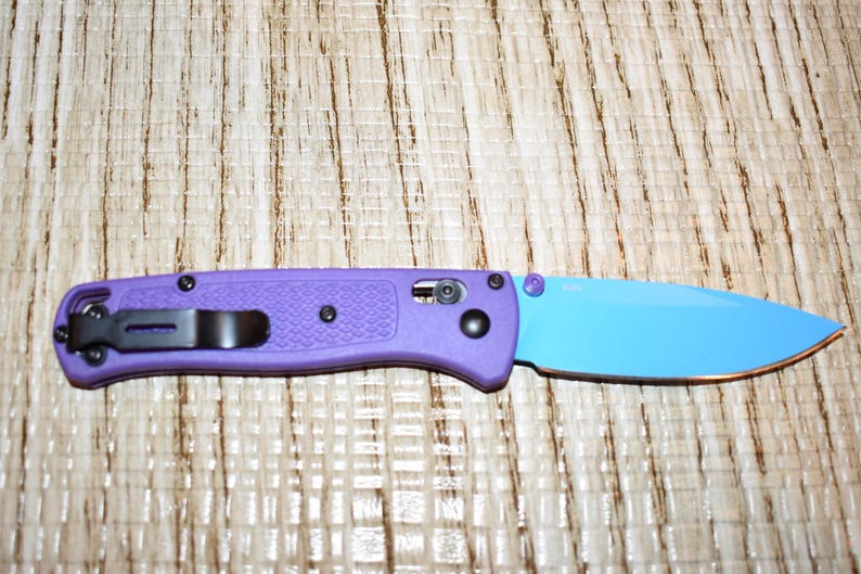 May include: A purple folding knife with a blue blade. The knife has a black pocket clip and black hardware. The handle is textured, and the blade is a solid blue color.