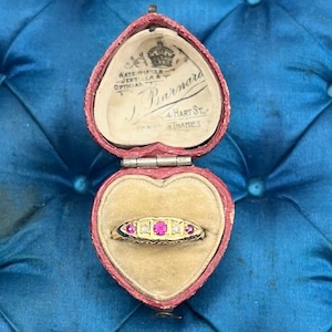 以下が含まれることがあります： 中央にピンク色の宝石、両側に2つのクリアストーンと2つの小さなピンク色の石を配したゴールドリング。赤いベルベットの裏地が付いたハート型の箱にセットされています。箱は開いており、クリーム色の内装にリングが映えます。箱は青色のキルティング加工された表面に置かれています。