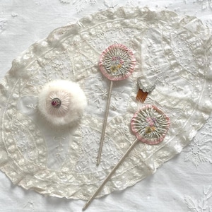 May include: An assortment of vintage vanity items, including a powder puff with a red jewel, two decorative flower-shaped items with pink and green accents, and a small hair comb. The items are arranged on a white lace doily.