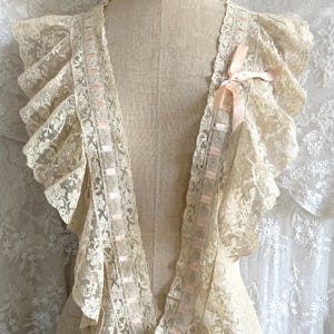 May include: Cream-colored lace collar with ruffled edges and pink ribbon accents. The delicate lace is draped over a mannequin, showcasing the intricate floral patterns and textures. A vintage-inspired accessory.