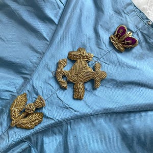 Peut inclure: Un vêtement bleu clair à fines rayures orné de trois écussons brodés dorés. L'écusson central représente une grande ancre, flanqué d'un emblème en forme de chardon sur la gauche et d'une couronne avec du velours violet sur la droite.
