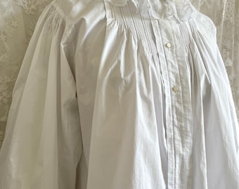 May include: A white blouse with a high, lace-trimmed collar and long, voluminous sleeves. The front features vertical pleats and small buttons. The cuffs are finished with lace. The blouse is displayed on a mannequin.