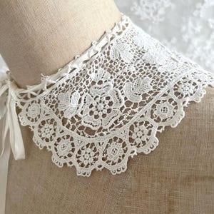May include: A white lace collar with a floral design. The collar is made of delicate, open-weave lace and features a scalloped edge. White satin ribbons are attached to the collar for tying. The collar is displayed on a mannequin.