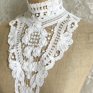 May include: A white lace collar featuring an intricate floral pattern with a high neck design. The lace is constructed with detailed embroidery and openwork, extending downward in a pointed shape. The piece is displayed on a beige mannequin.