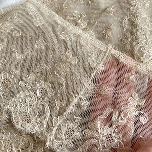 May include: Close-up of delicate, cream-colored lace fabric. The lace features intricate floral and scrollwork patterns, with a fine mesh background. The fabric is folded, revealing the detailed texture and design. The lace appears to be vintage or antique.