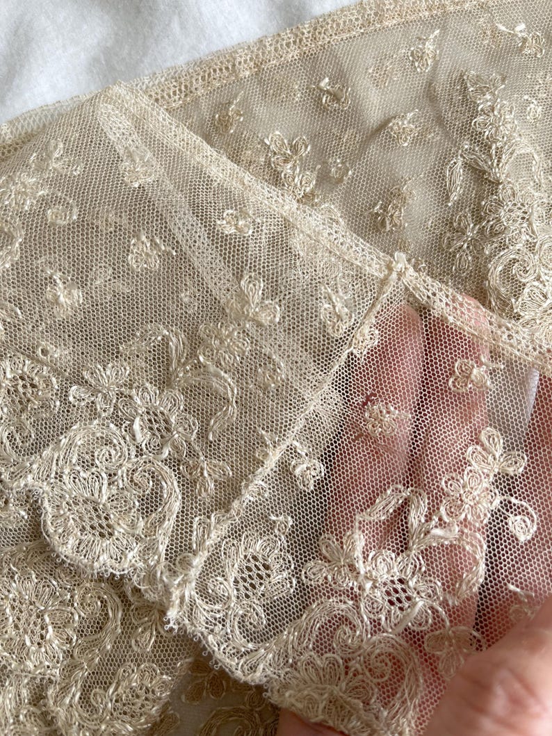 May include: Close-up of delicate, cream-colored lace fabric. The lace features intricate floral and scrollwork patterns, with a fine mesh background. The fabric is folded, revealing the detailed texture and design. The lace appears to be vintage or antique.