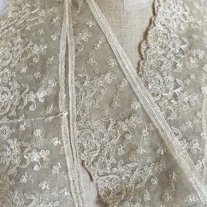 May include: A delicate, cream-colored lace shawl with intricate floral patterns. The shawl features a V-neck design and is draped over a mannequin. The lace is detailed with small, raised floral motifs and a scalloped edge.