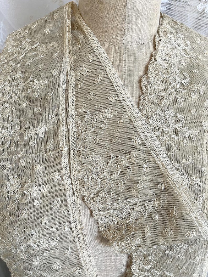 May include: A delicate, cream-colored lace shawl with intricate floral patterns. The shawl features a V-neck design and is draped over a mannequin. The lace is detailed with small, raised floral motifs and a scalloped edge.