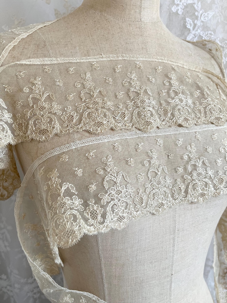 May include: Close-up of cream-colored lace fabric with floral and leaf patterns. The lace is delicate and detailed, with a scalloped edge. The fabric is draped over a mannequin, showcasing the texture and design of the lace.