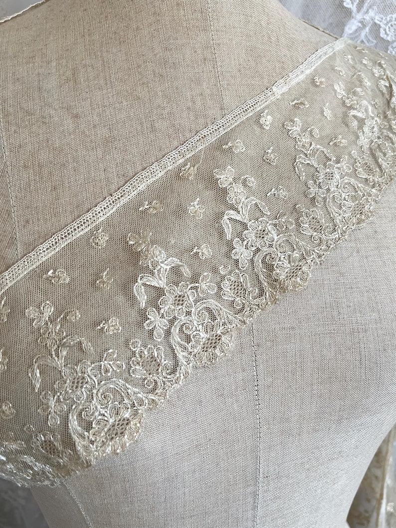 May include: Close-up of delicate ivory lace trim with floral and leaf patterns. The lace is intricately detailed with a scalloped edge and is displayed on a neutral-toned fabric background. The trim is likely used for embellishing garments or crafts.