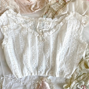 May include: A white, sleeveless lace top with a delicate, openwork design. The top features a ruffled neckline and small ribbon accents. Other vintage lace items are visible in the background, including a pink heart-shaped pillow.