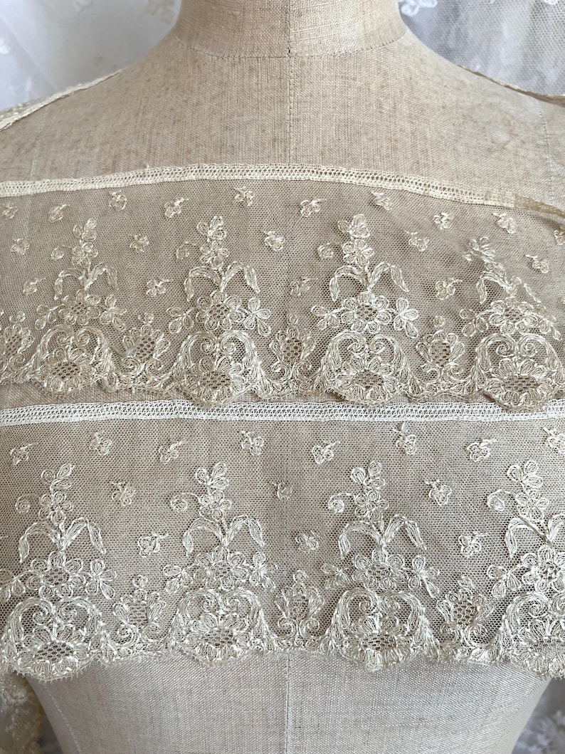 May include: Close-up of cream-colored lace fabric with floral and leaf patterns. The lace is detailed with delicate embroidery and a scalloped edge. The fabric is displayed on a mannequin.