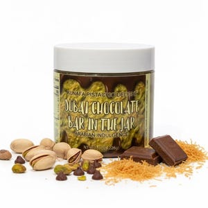 Puede incluir: Un tarro de "Dubai Chocolate Bar in the Jar" con el texto "Kunafa Pistachio Delight" y "Arabian Indulgence". El tarro está rodeado de pistachos, chispas de chocolate, repostería rallada y dos trozos de chocolate. Peso neto 226g.