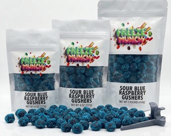 May include: Three white pouches of Sour Blue Raspberry Gushers candy, with the brand name "Freeze Munch" in colorful text. The pouches are labeled with the net weight: 4 oz, 8 oz, and 1 pound. A pile of blue candies and a gray scoop are in front.