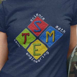 Puede incluir: Una camiseta azul marino con un gráfico de un diamante con las letras STEM en su interior. Las letras son de diferentes colores y cada letra está rodeada de un patrón diferente. Las palabras "ciencia", "tecnología", "ingeniería" y "matemáticas" están escritas alrededor del diamante.