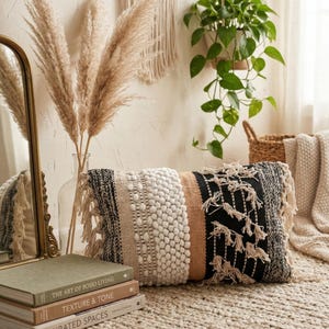 May include: A decorative lumbar pillow with a textured design. The pillow features a mix of black, white, and tan stripes with fringe detailing. The pillow is placed on a woven rug, with a stack of books and pampas grass in the background.