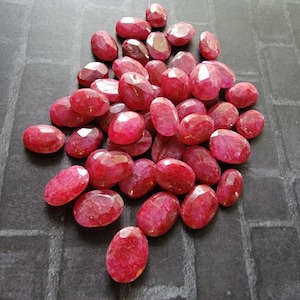 May include: A pile of oval-shaped, faceted ruby gemstones. The rubies are a deep red color and have a glossy finish.
