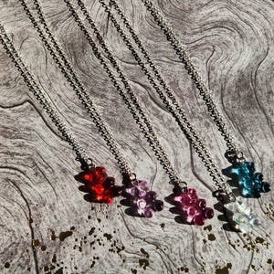 May include: Five silver chains with gummy bear charms in different colors: red, purple, pink, clear, and blue. The chains are hanging against a woodgrain background.